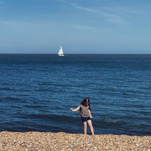 a little girl walks on Deal beach while in the background a sail boat goes by on the blue sea