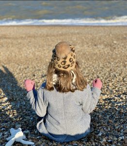 A little girl wearing a bobble hat sitting cross legged on Deal beach