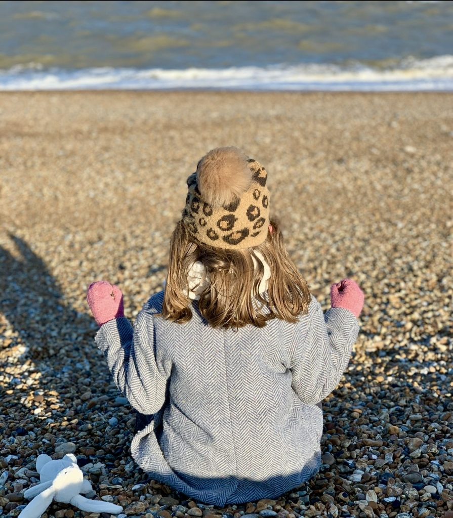 A little girl wearing a bobble hat sitting cross legged on Deal beach